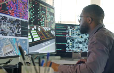 Engineer reviewing circuit board schematics and technical diagrams across multiple monitors