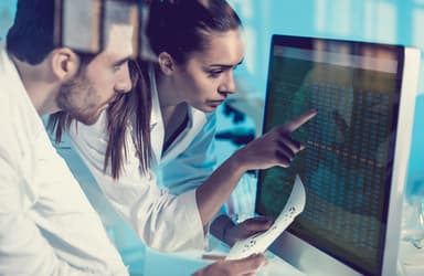 Two researchers in lab coats reviewing data charts on a monitor, relevant to digital and data policy in the public sector